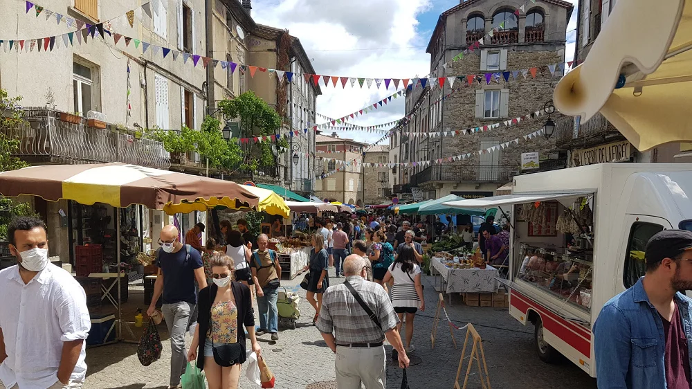 Le marché des Vans, un incontournable des marchés en Ardèche Foule déambulant parmi les étals du marché des Vans, l’un des marchés les plus vivants d’Ardèche, sous des fanions colorés.