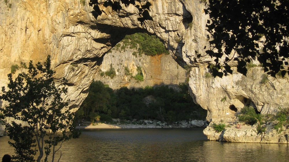 Pont d'Arc, une grande arche naturelle en pierre calcaire, illuminée par le soleil couchant. La rivière Ardèche coule en dessous, et la plage du Pont d'Arc est visible en arrière-plan.