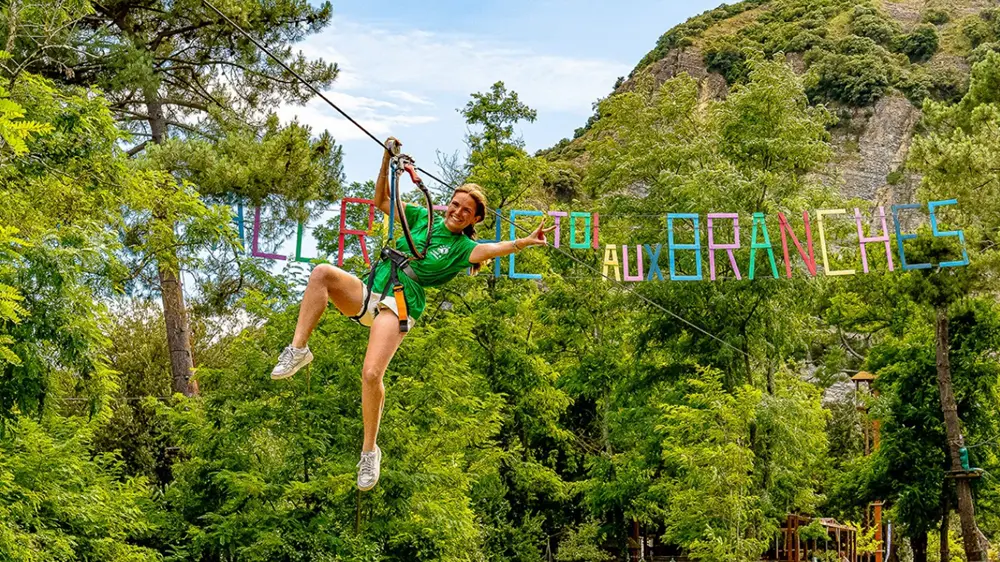 Accroche-toi aux branches 34 Femme souriante en harnais sur une tyrolienne dans un parc accrobranche entouré d’arbres verts et d’une banderole colorée "Accroche toi aux Branches".