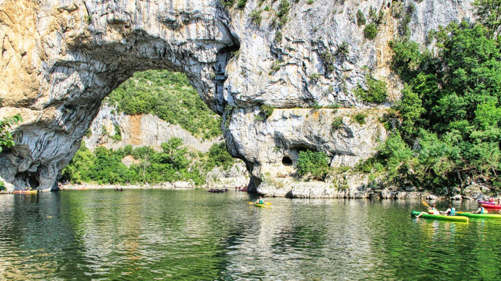 Rivière Ardèche et canoës sous le Pont d’Arc – baignade en Ardèche Rivière Ardèche sous le Pont d’Arc avec des canoës, spot populaire pour baignade en Ardèche et activités nautiques.