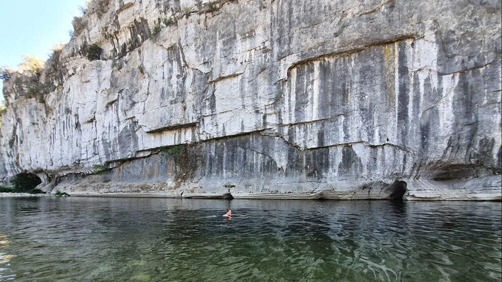 Nage à la Padelle baignade, falaises majestueuses en Ardèche Femme nageant dans l’eau profonde de la Padelle baignade, avec de hautes falaises calcaires en arrière-plan.