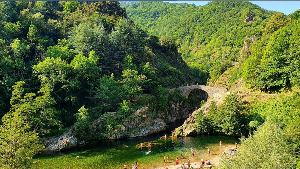 Le Pont du Diable à Thueyts : nature, baignade et patrimoine en Ardèche Vue estivale du Pont du Diable en Ardèche, à Thueyts, avec des baigneurs dans la rivière et la forêt verdoyante en arrière-plan