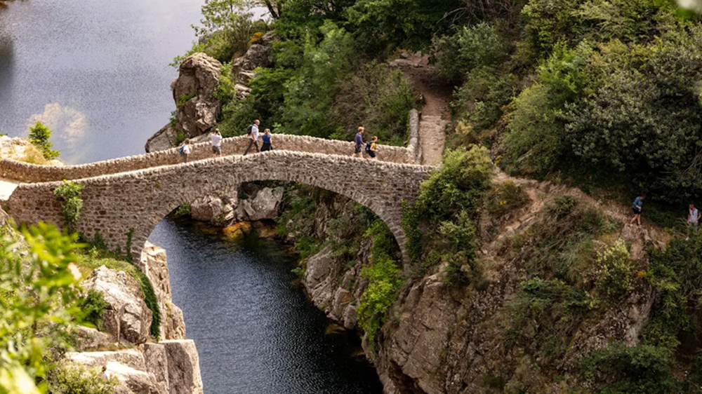 Baignade au pied du Pont du Diable à Thueyts : un écrin naturel en Ardèche Vue du Pont du Diable à Thueyts avec des baigneurs dans la rivière Ardèche, entouré de falaises verdoyantes et de rochers