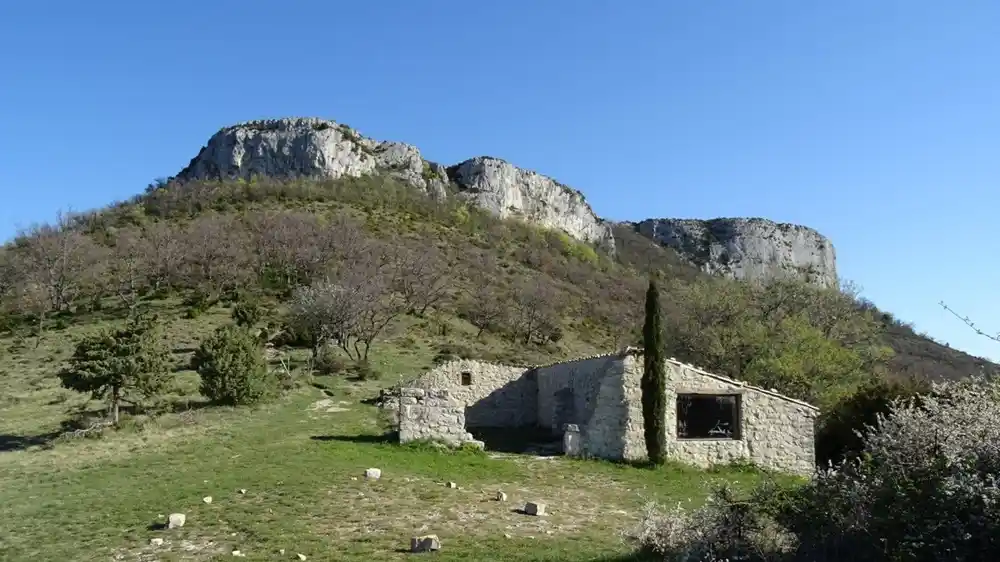 dent-de-rez-sud-ardeche-paysage-ruine-pierre Vue en perspective depuis une pente herbeuse, montrant une ancienne ruine de bâtisse en pierre calcaire restaurée avec une fenêtre carrée. Un cyprès solitaire se tient à côté de la ruine. En arrière-plan, l'imposante masse rocheuse calcaire (la Dent de Rez) domine le paysage sous un ciel bleu clair, couverte de végétation arbustive.