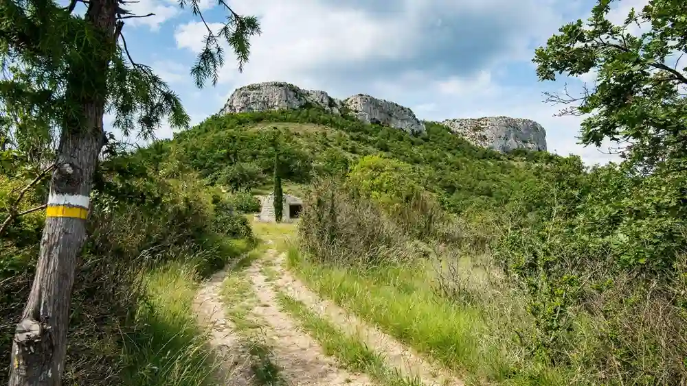 sentier-randonnee-vers-dent-de-rez-ardeche Vue en perspective d'un sentier de terre s'élevant à travers la garrigue dense. Un marquage de randonnée jaune et blanc est visible sur un arbre au premier plan à gauche. Le sentier mène vers une petite cabane en pierre (abri ou petite chapelle) et, en arrière-plan, l'imposante masse rocheuse calcaire du sommet de la Dent de Rez sous un ciel nuageux.