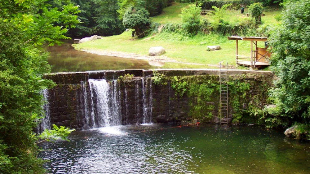 espace de baignade la neuve 2 Vue d'ensemble d'une zone de baignade naturelle à l'espace de baignade la neuve, avec un vieux barrage en pierre formant une cascade dans une piscine naturelle profonde. Rive en herbe et forêt en arrière-plan près de Privas.