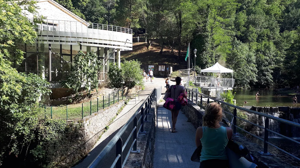 espace de baignade la neuve 3 Entrée de l'espace de baignade la neuve avec deux personnes marchant sur une passerelle. Le bâtiment des installations est à gauche et la zone de baignade avec cascade près de Privas est à droite, entourée de forêts.