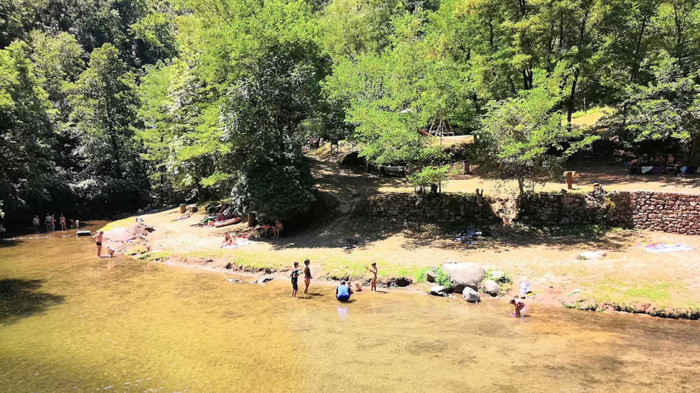 La plage animée et sécurisée de l’espace de baignade la neuve : le lieu incontournable pour une baignade à privas rafraîchissante et en toute sérénité en famille. Entrée de l'espace de baignade la neuve avec deux personnes marchant sur une passerelle. Le bâtiment des installations est à gauche et la zone de baignade avec cascade près de Privas est à droite, entourée de forêts.