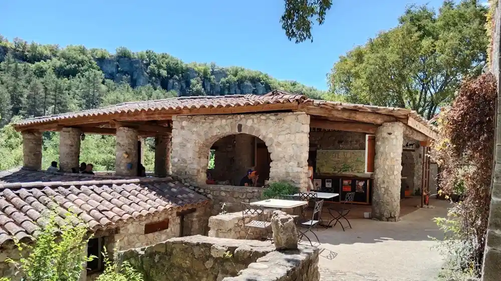 Photo d'un bâtiment traditionnel en pierre au Vieil Audon, avec terrasse couverte, arches et piliers en pierre. Des personnes sont assises à des tables de café sous un toit en tuiles de terre cuite. En arrière-plan, une falaise calcaire boisée. Le site est entouré de végétation.