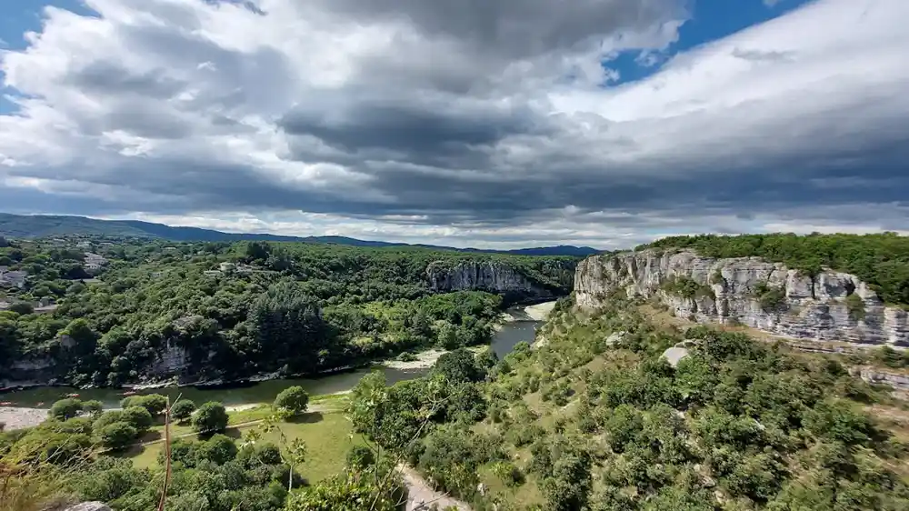 Photo panoramique d'un paysage de gorges avec la rivière Ardèche serpentant entre des falaises calcaires boisées. La vue est prise depuis un point de vue élevé (tour Jeanne), donnant sur la vallée verte et le Vieil Audon. Ciel bleu avec des nuages orageux.