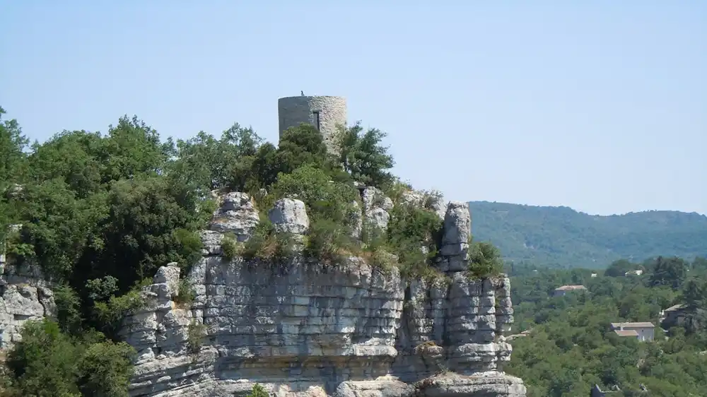Photo d'une tour médiévale circulaire construite en pierres locales claires, située au sommet d'un haut éperon rocheux calcaire. La tour est entourée d'arbres verts et de végétation dense. En arrière-plan, on distingue la vallée et d'autres collines sous un ciel bleu clair.